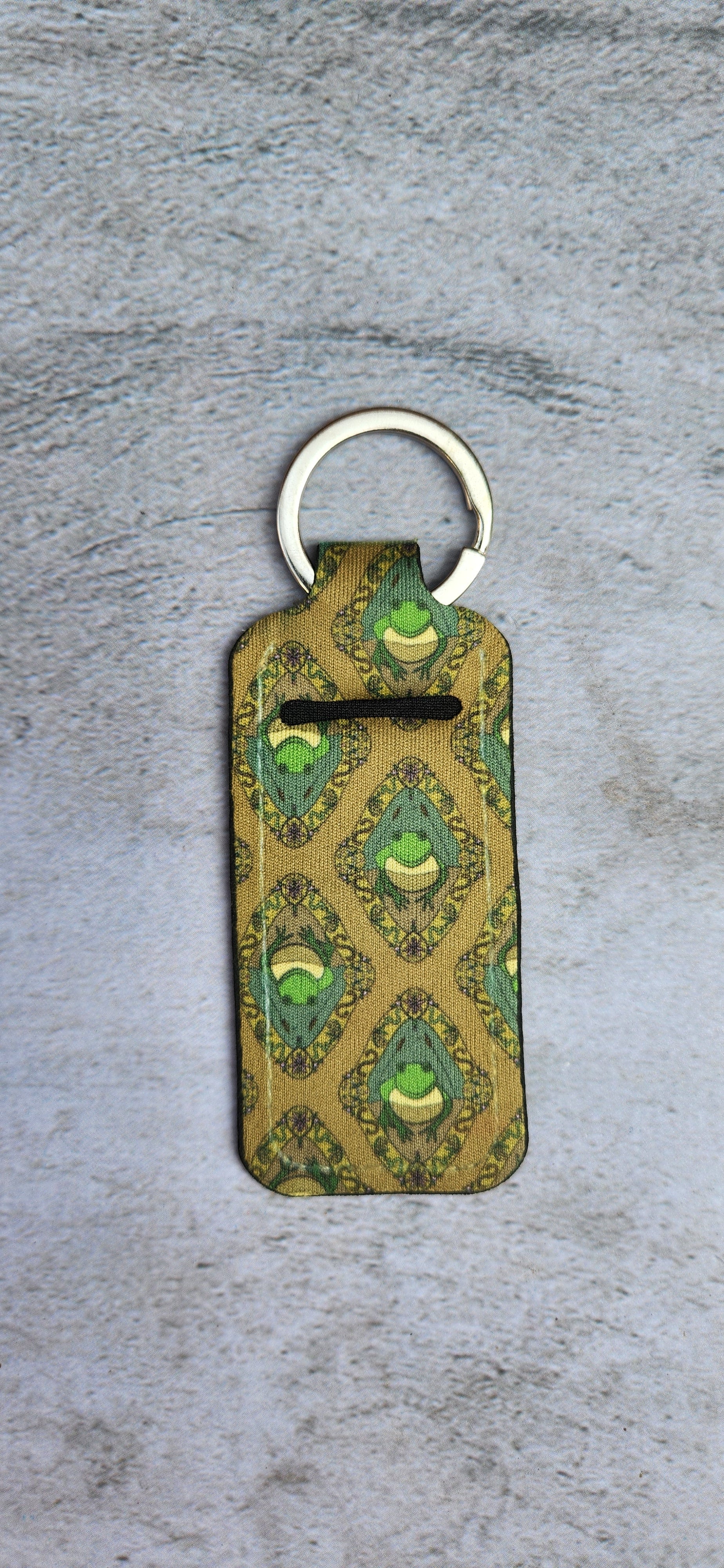 Lip Balm key chain with green and gold frog pattern on concrete background